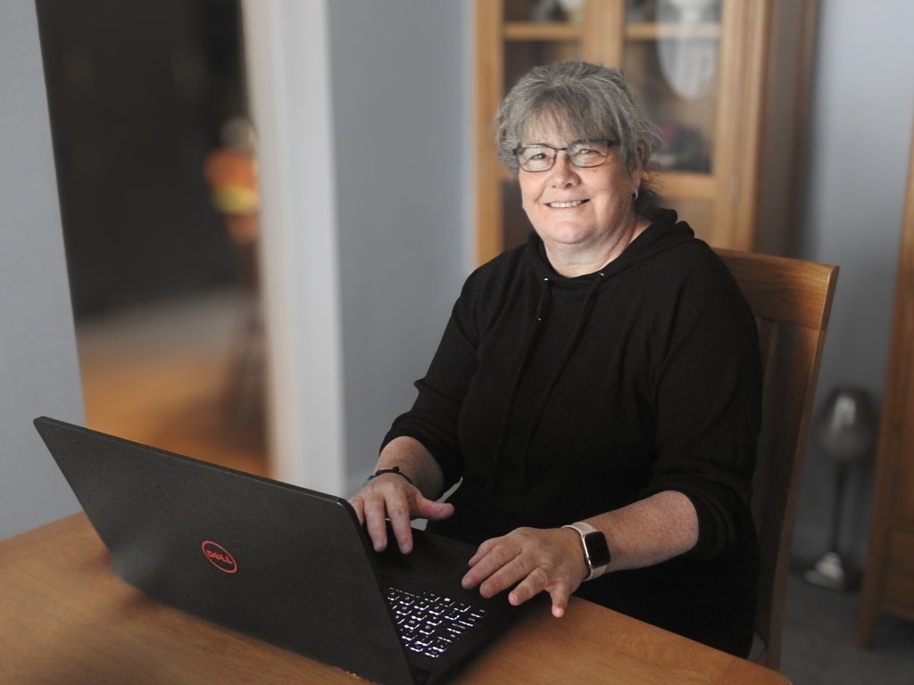Debbie Walton from Mindful Growth Solutions sitting at a wooden table using a laptop, wearing glasses and a black hoodie, smiling at the camera.