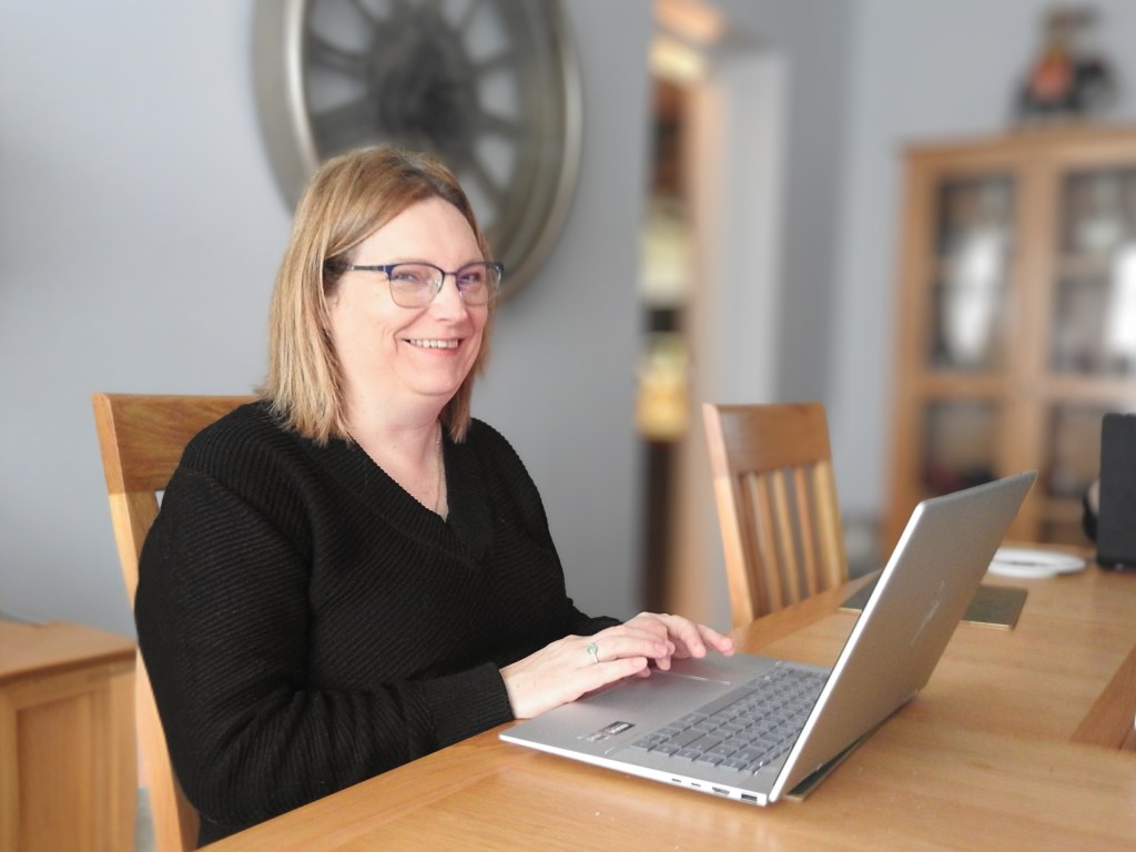 Angela Bird from Mindful Growth Solutions sits at a wooden table using a laptop, smiling at the camera.